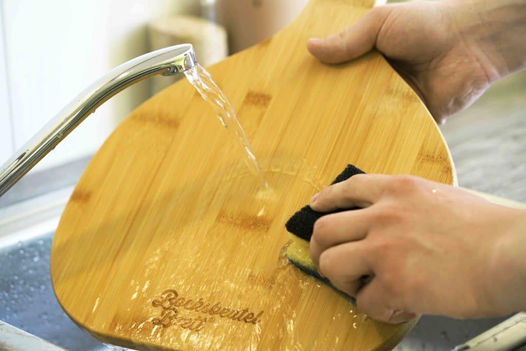 Ein Küchenbrett aus Holz wird unter fließendem Wasser per Hand gespült.